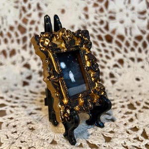 May include: A miniature gold-framed picture with a black background and white shapes inside, displayed on a black easel.