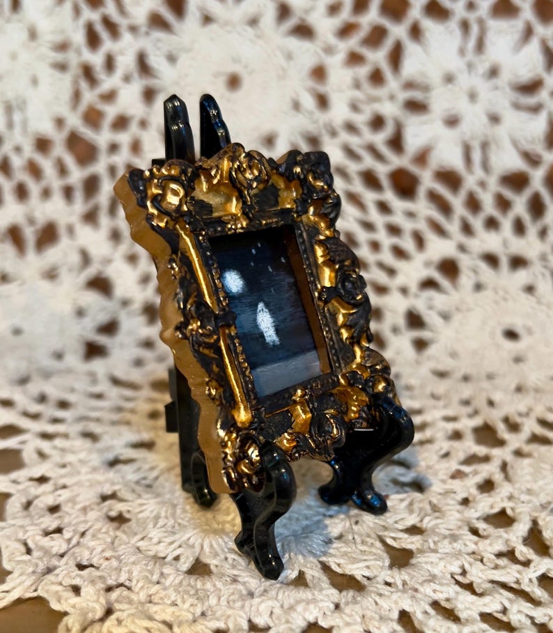 May include: A miniature gold-framed picture with a black background and white shapes inside, displayed on a black easel.