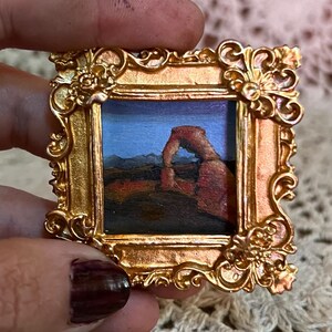 May include: Miniature painting of a desert landscape with a natural arch, set within a small, ornate gold frame. The painting features a blue sky, brown rock formations, and dark ground. The frame has intricate floral and scroll details.