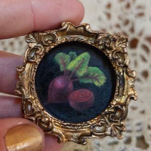 May include: A small, ornate gold-colored frame surrounds a circular painting of a beet with green and red leaves. The painting is set against a dark background. The frame has a detailed, vintage design.