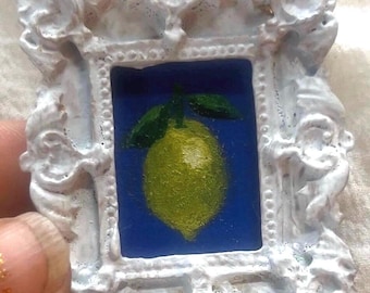 May include: A small white ornate frame with a painting of a yellow lemon with green leaves on a blue background.