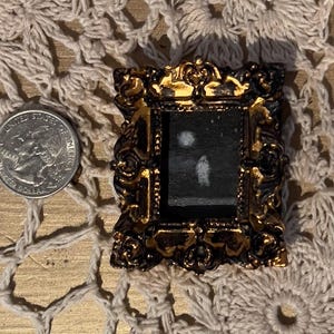 May include: A small, ornate gold-colored frame with a black background and a white silhouette of a person inside. The frame is sitting on a white crocheted surface.