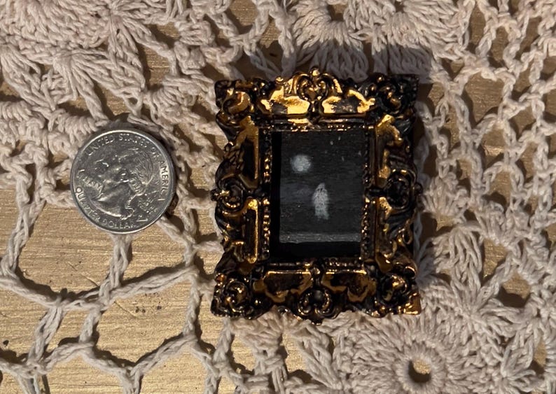 May include: A small, ornate gold-colored frame with a black background and a white silhouette of a person inside. The frame is sitting on a white crocheted surface.