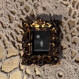 May include: A small, ornate gold frame with a black background and a white silhouette of a person standing in front of a white circle.