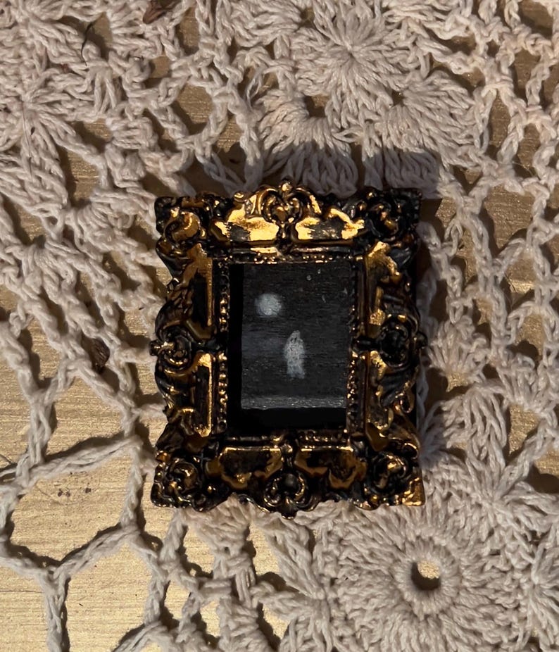 May include: A small, ornate gold frame with a black background and a white silhouette of a person standing in front of a white circle.