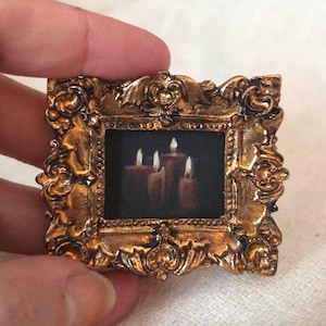 May include: A miniature painting of lit candles in a dark room, set within an ornate, gold-colored frame. The painting features three candles with flickering flames, creating a warm, inviting glow. The frame has a detailed, baroque design.
