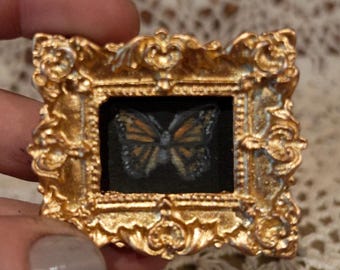 May include: A small, ornate gold frame with a black background and a painted image of a butterfly with orange and black wings.