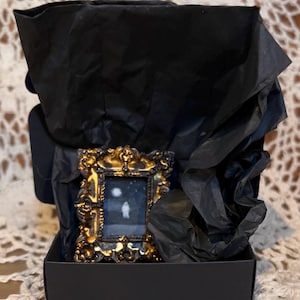 May include: A small, ornate gold frame with a black background and a white silhouette of a person inside. The frame is sitting in a black box with black tissue paper.