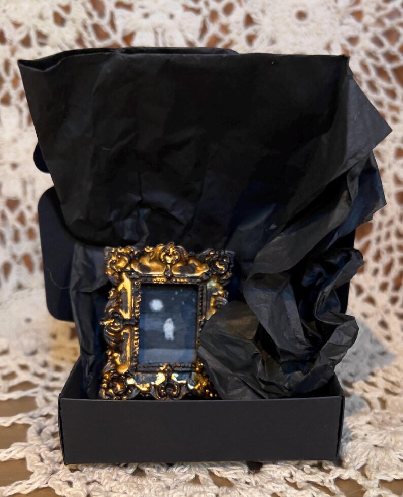 May include: A small, ornate gold frame with a black background and a white silhouette of a person inside. The frame is sitting in a black box with black tissue paper.