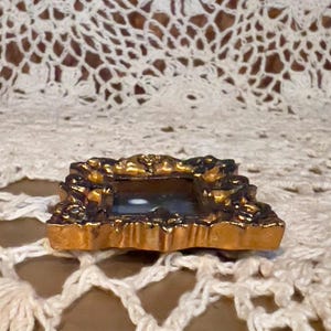 May include: A small, ornate gold-colored picture frame with a dark interior. The frame has a detailed, decorative design.