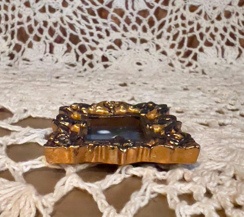 May include: A small, ornate gold-colored picture frame with a dark interior. The frame has a detailed, decorative design.