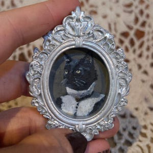 May include: A miniature portrait of a black cat with yellow eyes, wearing a white collar, set within an ornate silver-colored frame. The oval painting has a vintage aesthetic, reminiscent of classic portraiture.