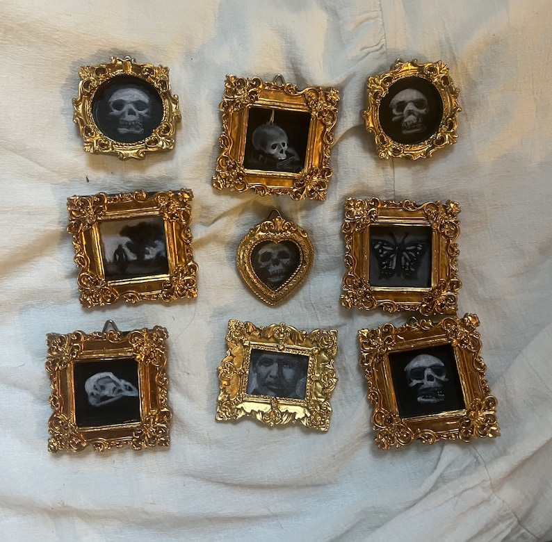 May include: Nine gold-toned picture frames with ornate designs, each containing a black and white image of a skull, butterfly, or other design. The frames are arranged in a grid pattern on a white fabric surface.