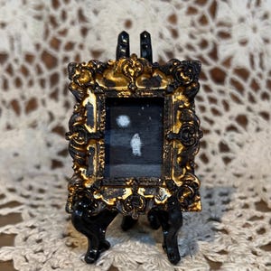 May include: A miniature painting of a snowy landscape with two white circles, possibly representing the moon and a distant light, framed in a gold ornate frame and displayed on a black easel.