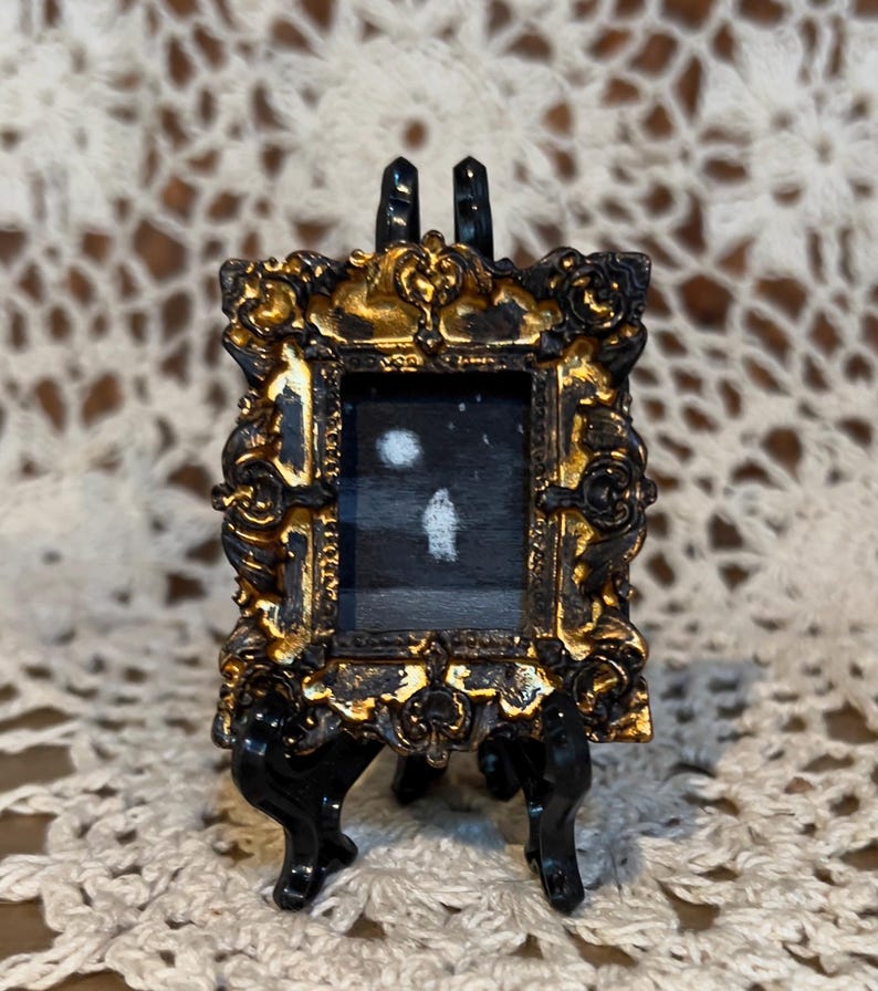 May include: A miniature painting of a snowy landscape with two white circles, possibly representing the moon and a distant light, framed in a gold ornate frame and displayed on a black easel.
