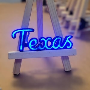 May include: A blue neon sign that spells out the word "Texas". The sign is displayed on a small wooden easel. The neon light gives off a bright blue glow, making the word stand out.