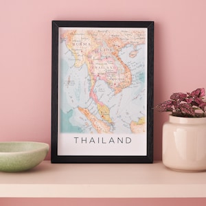 Könnte beinhalten: Eine gerahmte Vintage-Karte von Thailand mit einem schwarzen Rahmen. Die Karte ist auf weißem Papier mit blauen, grünen und rosa Farben gedruckt. Die Karte zeigt die umliegenden Länder Burma, Laos, Kambodscha und Malaysia. Der Text "THAILAND" ist in Schwarz am unteren Rand der Karte gedruckt.