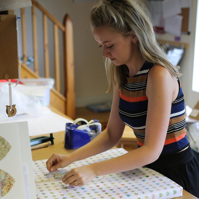 May include: A person wrapping a gift with patterned paper. The person is wearing a striped top with orange, blue, and brown stripes. The gift is wrapped on a wooden table with tape and other supplies nearby.