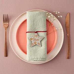 May include: A light green linen napkin with a red ribbon tied around it. A wooden star with the name "Oliver" carved into it is attached to the ribbon. The napkin is placed on a white and coral dinner plate.