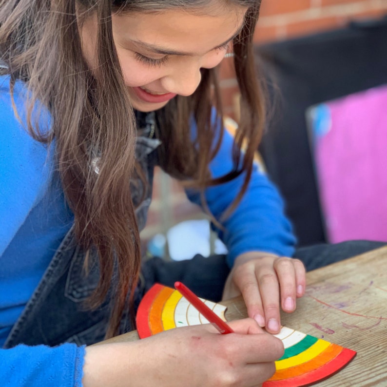 May include: A young person is coloring a wooden rainbow with a red crayon. The rainbow is made of multiple colors, including red, orange, yellow, green, blue, and purple. The rainbow is on a wooden surface.