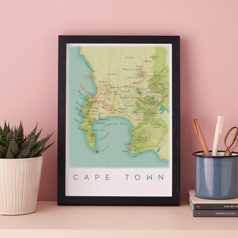 May include: A framed map print of Cape Town, South Africa, with a black frame. The map features a detailed illustration of the city's layout, including roads, landmarks, and the surrounding coastline. The words "CAPE TOWN" are printed in white at the bottom.