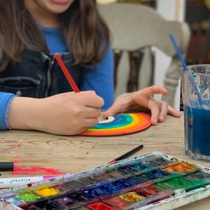 May include: A child paints a rainbow-colored wooden craft with a red paintbrush. A watercolor palette with various colors is on the table.