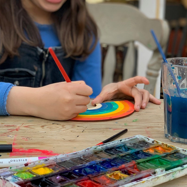 May include: A child paints a rainbow-colored wooden craft with a red paintbrush. A watercolor palette with various colors is on the table.