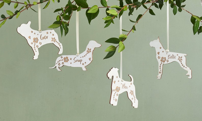 May include: Four white wooden dog ornaments with floral designs and names "Coco", "Snap", "Max", and "Bella" hanging from white ribbon on a green background.