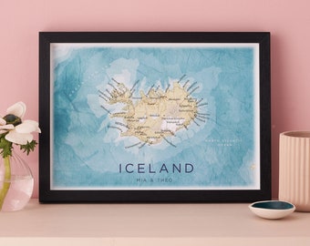 May include: A framed map of Iceland with a watercolor background. The map is in a black frame and the title "Iceland" is printed in navy blue with the names "Mia & Theo" underneath. The map shows the outline of Iceland with the names of cities and towns printed in black.
