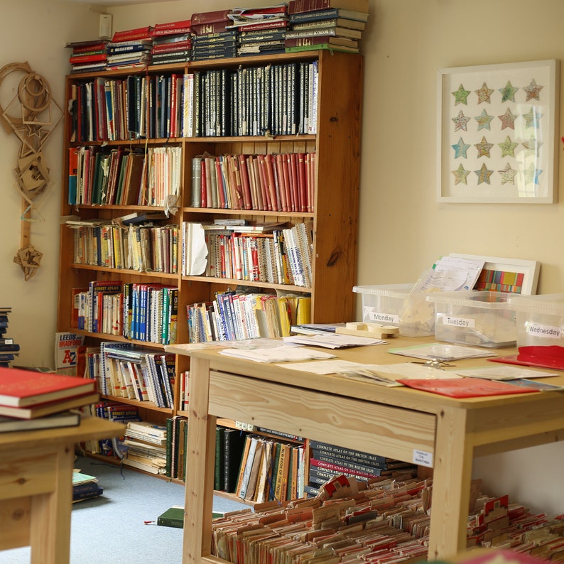 May include: A home office or library with a large wooden bookshelf filled with books of various sizes and colors. A wooden desk with papers and labeled storage bins sits in front of the bookshelf. A framed star-shaped artwork hangs on the wall.