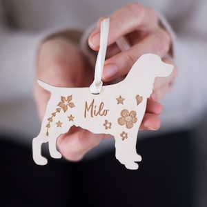 May include: A wooden dog ornament with the name "Milo" engraved on it. The ornament is decorated with flowers and stars.