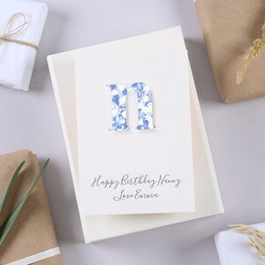 May include: A white card with a blue and white floral letter "N" and the text "Happy Birthday Nancy Love Emma".