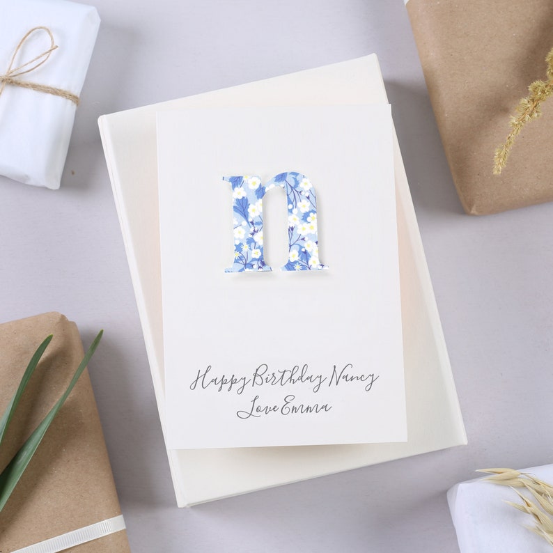 May include: A white card with a blue and white floral letter "N" and the text "Happy Birthday Nancy Love Emma".