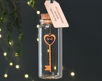 May include: A clear glass vial ornament with a cork stopper containing a wooden key cutout with a heart-shaped bow. A wooden tag hangs from a teal ribbon, engraved with the text 1ST CHRISTMAS IN OUR NEW HOME, suspended by a thin string.