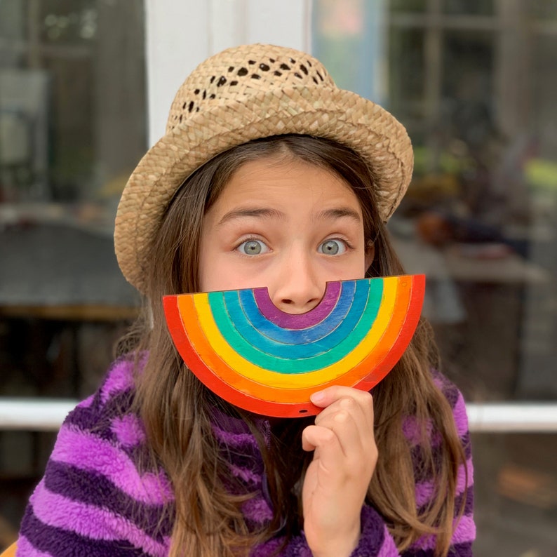May include: A young person wearing a straw hat holds a rainbow-colored wooden half-circle in front of their face.