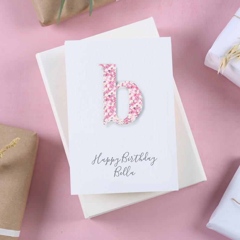 May include: A white card with a pink floral letter "b" and the text "Happy Birthday Bella".