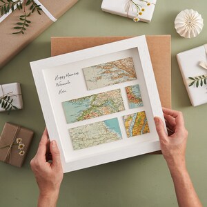 May include: A white framed artwork featuring vintage map pieces. The artwork includes the handwritten text "Happy Memories Worldwide Here." The maps are in shades of blue, green, and tan. The background includes wrapped gifts and decorative elements.