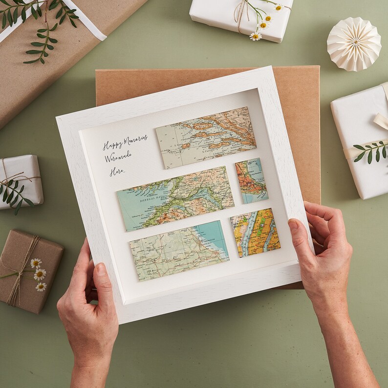 May include: A white framed artwork featuring vintage map pieces. The artwork includes the handwritten text "Happy Memories Worldwide Here." The maps are in shades of blue, green, and tan. The background includes wrapped gifts and decorative elements.