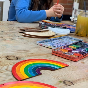 May include: Two rainbow-colored wooden arches painted with watercolors. The arches are on a wooden table with a paint palette and paintbrushes.