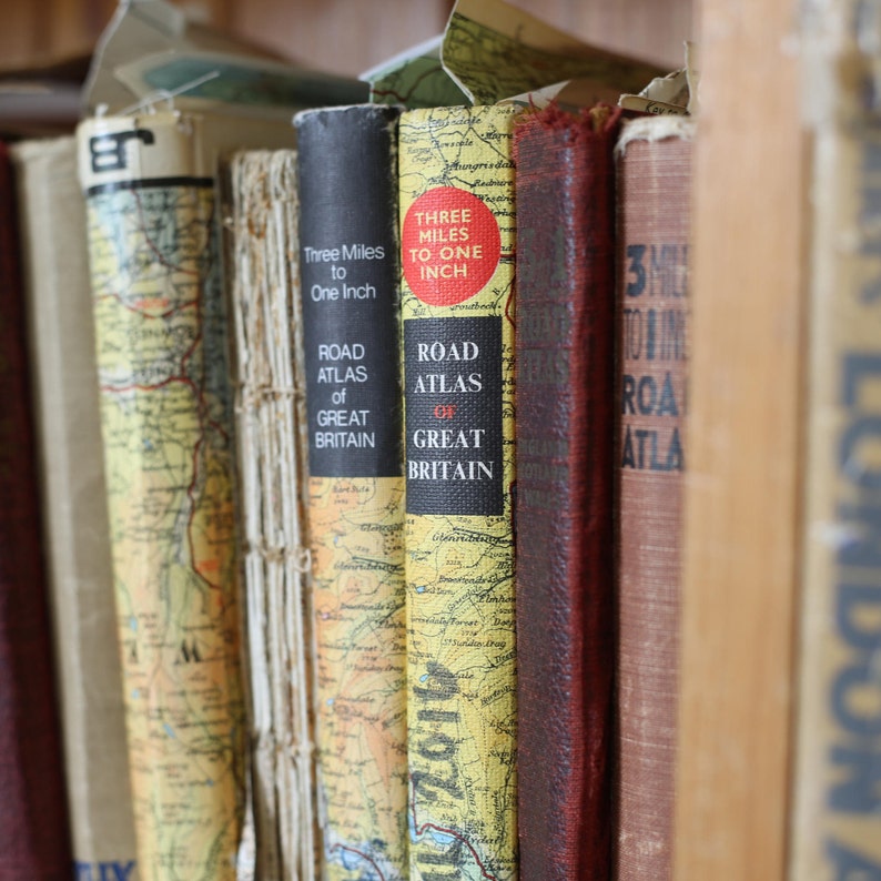 May include: A collection of vintage road atlases of Great Britain, with titles like "Three Miles to One Inch." The spines are a mix of colors, including yellow, black, and red, with map designs. The books are arranged on a shelf.