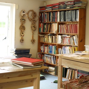 May include: A room with wooden bookshelves filled with books of various sizes and colors. A wooden table in the foreground holds a stack of books with a red cover. Decorative paper art hangs on the wall.