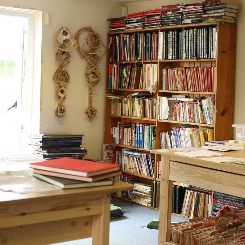 May include: A room with wooden bookshelves filled with books of various sizes and colors. A wooden table in the foreground holds a stack of books with a red cover. Decorative paper art hangs on the wall.