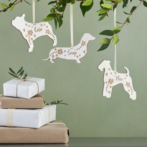 May include: Three white wooden dog ornaments with floral details and personalized names: Coco, Snoop, and Max. The ornaments are hanging from white ribbon on a green background.