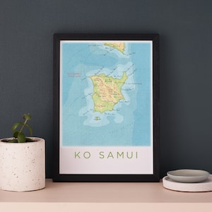 Könnte beinhalten: Ein gerahmter Druck einer Karte von Ko Samui, Thailand. Die Karte ist im Vintage-Stil mit blauem Hintergrund und grünen Landmassen gehalten. Der Text "KO SAMUI" ist in Grün unter der Karte gedruckt.