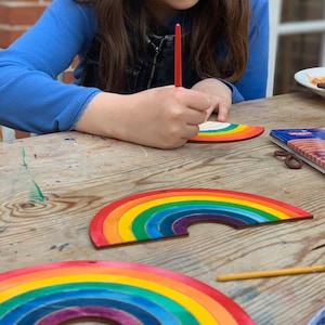 May include: A child paints a wooden rainbow craft with red paint. The rainbow is made of multiple layers of wood painted in different colors. The rainbow is on a wooden table.