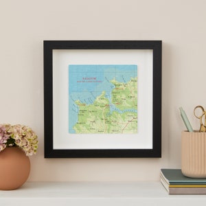 May include: A framed map print of Padstow and the Camel Estuary, featuring a detailed topographical view with green, blue, and brown hues. The print is set within a black frame and white matting, displayed on a white surface.
