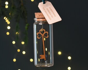 May include: A clear glass vial with a cork stopper containing a wooden key cutout with a clover-shaped bow. A rectangular wooden tag hangs from a dark green ribbon, engraved with the text CHRISTMAS WISHES TO THE SAUNDERS in capital letters.