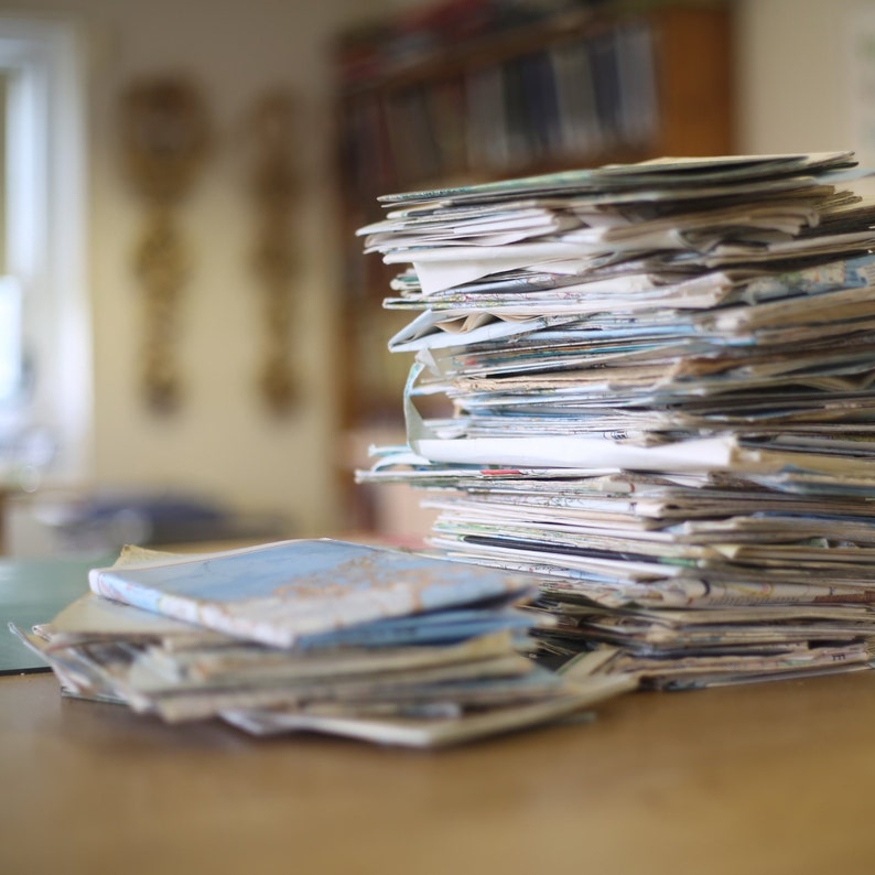 May include: A large stack of assorted papers and documents, including maps, sits on a wooden surface. The papers vary in size and color, with some featuring blue and white designs. The background is blurred, suggesting an indoor setting.