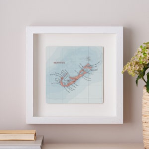 May include: A white framed map of Bermuda with a light blue background and pink and brown lines showing the island's shape and coastline. The map is titled "Bermuda" in the top left corner.