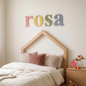 Puede incluir: Una escena de dormitorio con un cabecero de madera en forma de casa sobre una cama con almohadas y una colcha. Encima del cabecero, el nombre "rosa" está escrito con letras de madera de colores pastel. Una mesita de noche de madera contiene una flor y una decoración en forma de estrella.